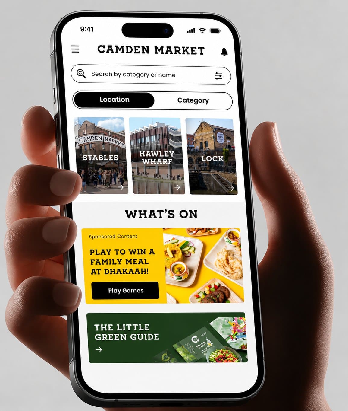 Hand holding a phone showing the Camden Market app home screen with location tabs for Stables, Hawley Wharf and Lock, a What's On sponsored game and The Little Green Guide, on a light grey background