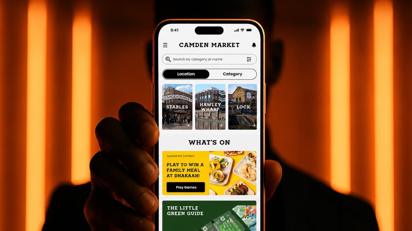 Silhouetted hand holding a phone showing the Camden Market app home screen with Stables, Hawley Wharf and Lock locations and a What's On promotion, against glowing orange light bars