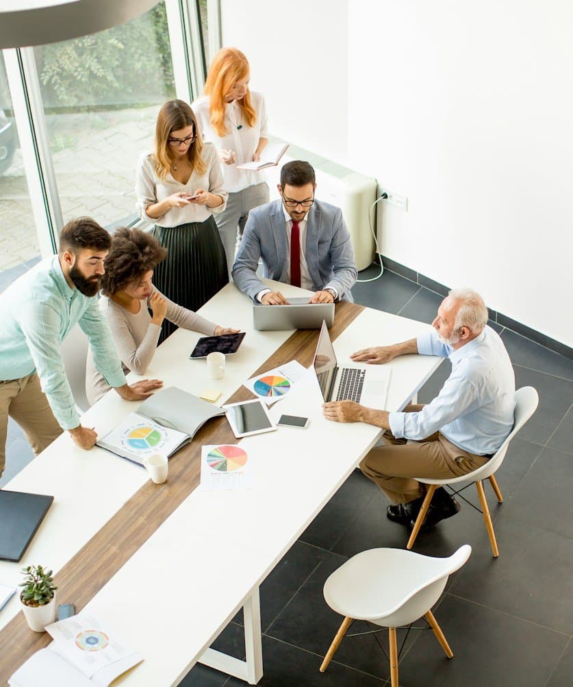 Overhead view of a product team collaborating around a white meeting table with laptops, tablets and printed charts in a bright office
