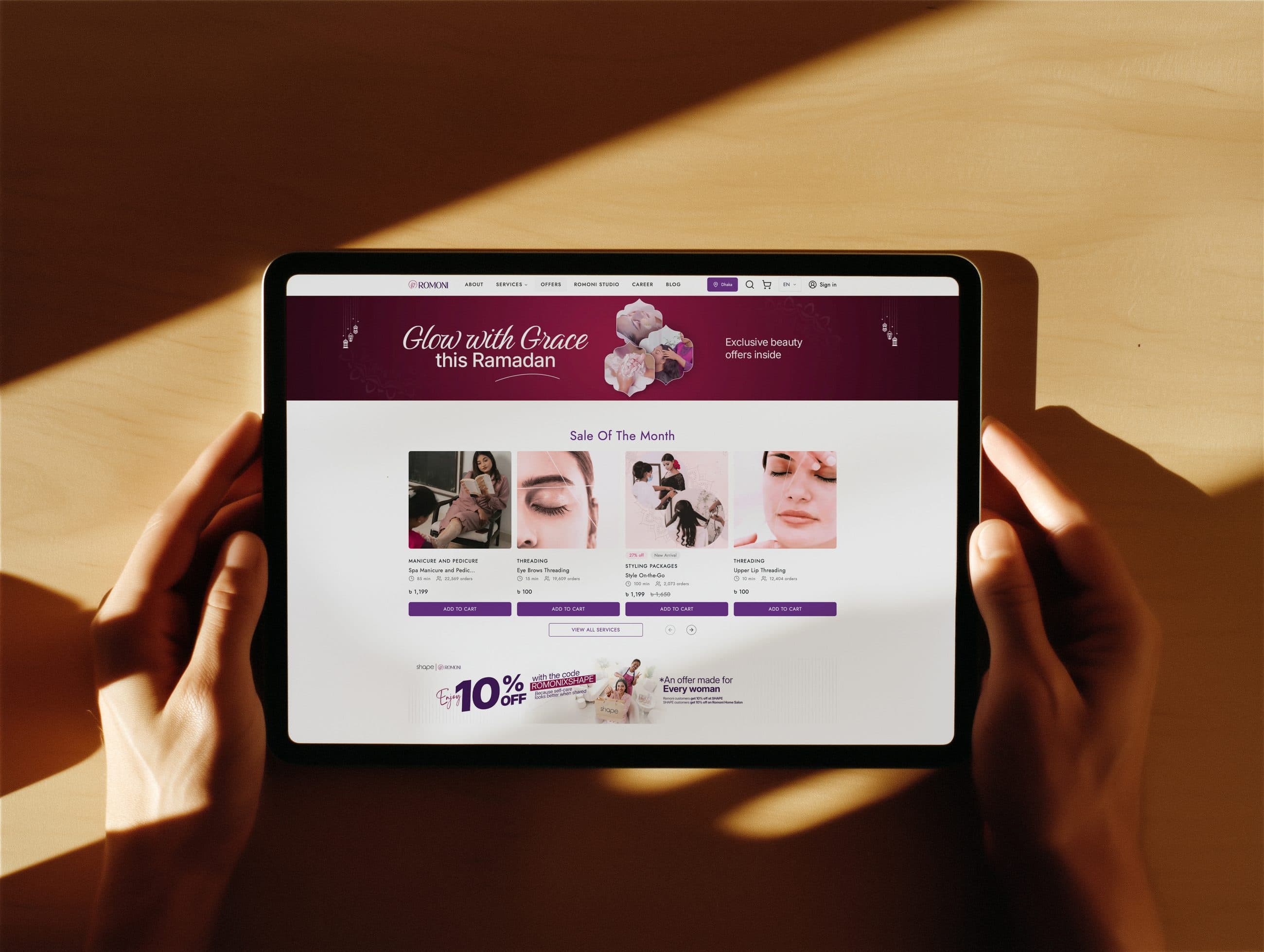 Two hands holding a tablet showing the Romoni website with a Ramadan campaign banner and the Sale of the Month services.