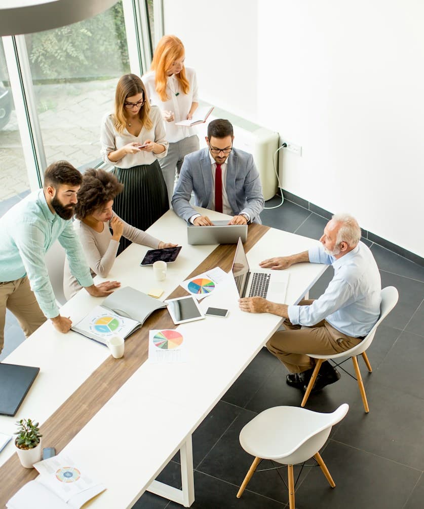 A product team gathered around a meeting table with laptops, tablets and printed charts.